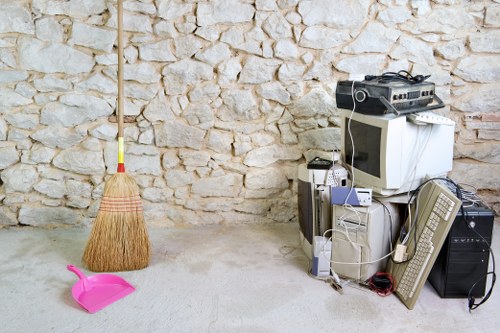 The image shows a collection of discarded household items placed on a light-colored concrete floor against a textured stone wall. In the foreground, there is a bright pink plastic dustpan with a matching handle, and a yellow and red broom with natural straw bristles positioned upright nearby. To the right, an assortment of electronic waste is stacked, including an old computer monitor with a bulky CRT screen, a tower CPU, and several boxed or loose cables and power supplies. An air conditioning remote control and various other small electronic components are also visible among the clutter. The environment suggests a domestic or backyard setting, possibly prepared for rubbish removal, with the electronic waste indicating content targeted for specialist disposal services such as those provided by Commercial Waste Removal Stockwell. The overall scene depicts typical household waste awaiting collection or removal, with a focus on bulky electronic items and cleaning tools, consistent with services around the Stockwell area post code SW9. This setting emphasizes the importance of proper waste separation and disposal for household electronics and general rubbish.