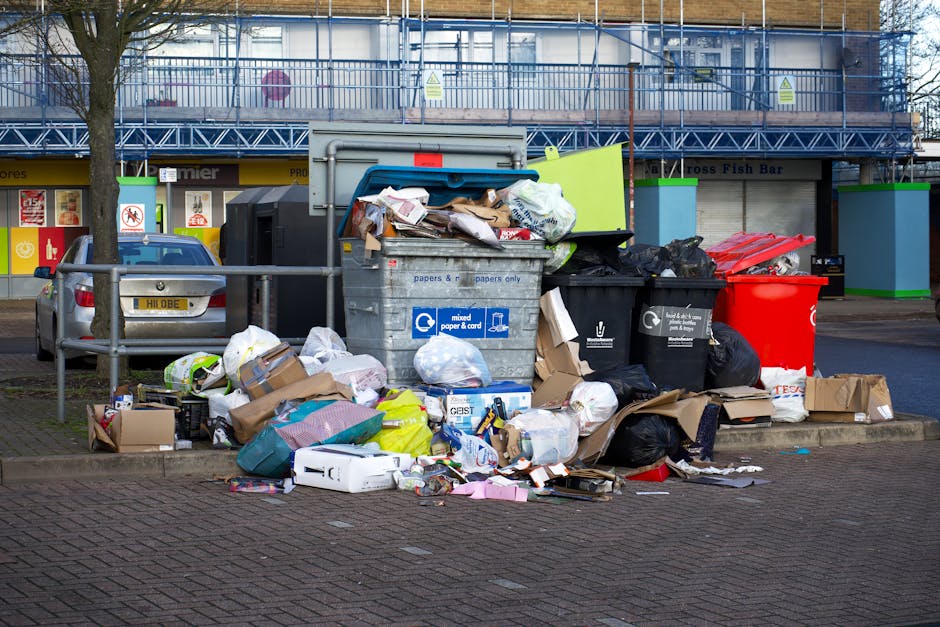 A worker wearing a high-visibility red and yellow jacket is attending to a large, red garbage collection truck parked on the side of a street. The truck's rear loading mechanism is open, revealing its
