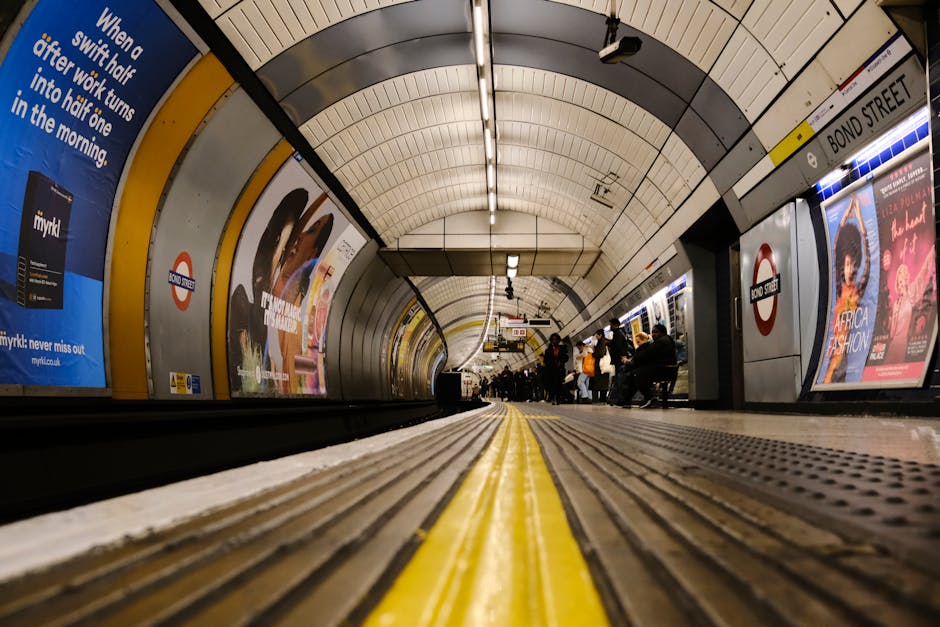 A photograph of an underground train station platform showing the curved tunnel entrance in the background. The platform is lit with bright overhead lights, casting reflections on the polished black f