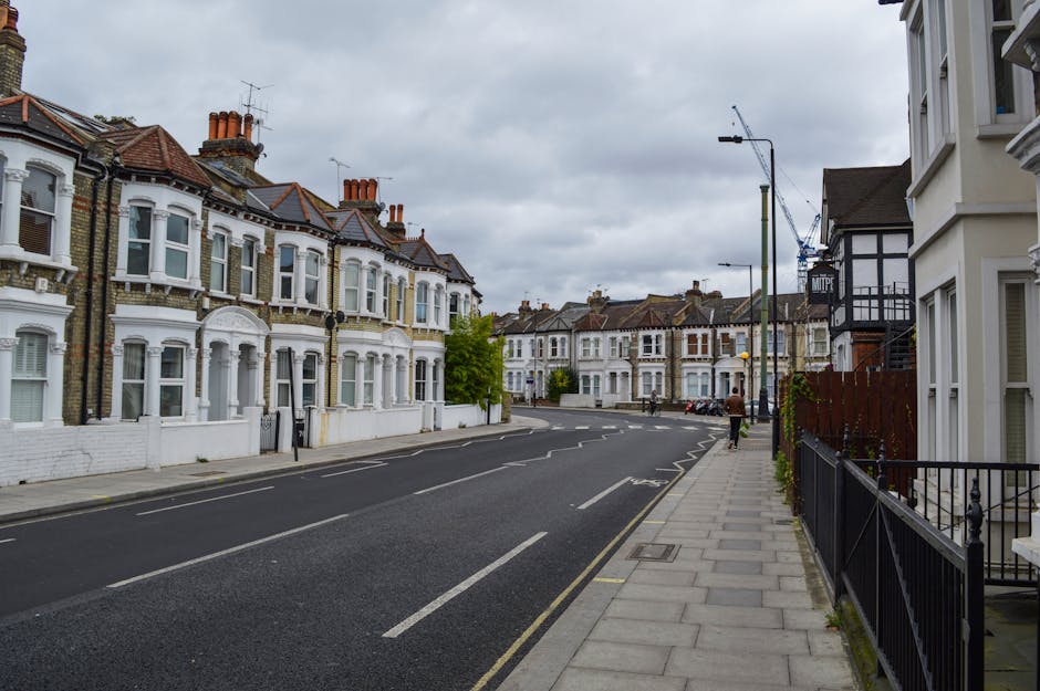 Piles of mixed waste, including cardboard boxes, plastic bags, paper, and packaging materials, are overflowing from large grey, black, and red wheelie bins situated on a paved urban street. The grey b